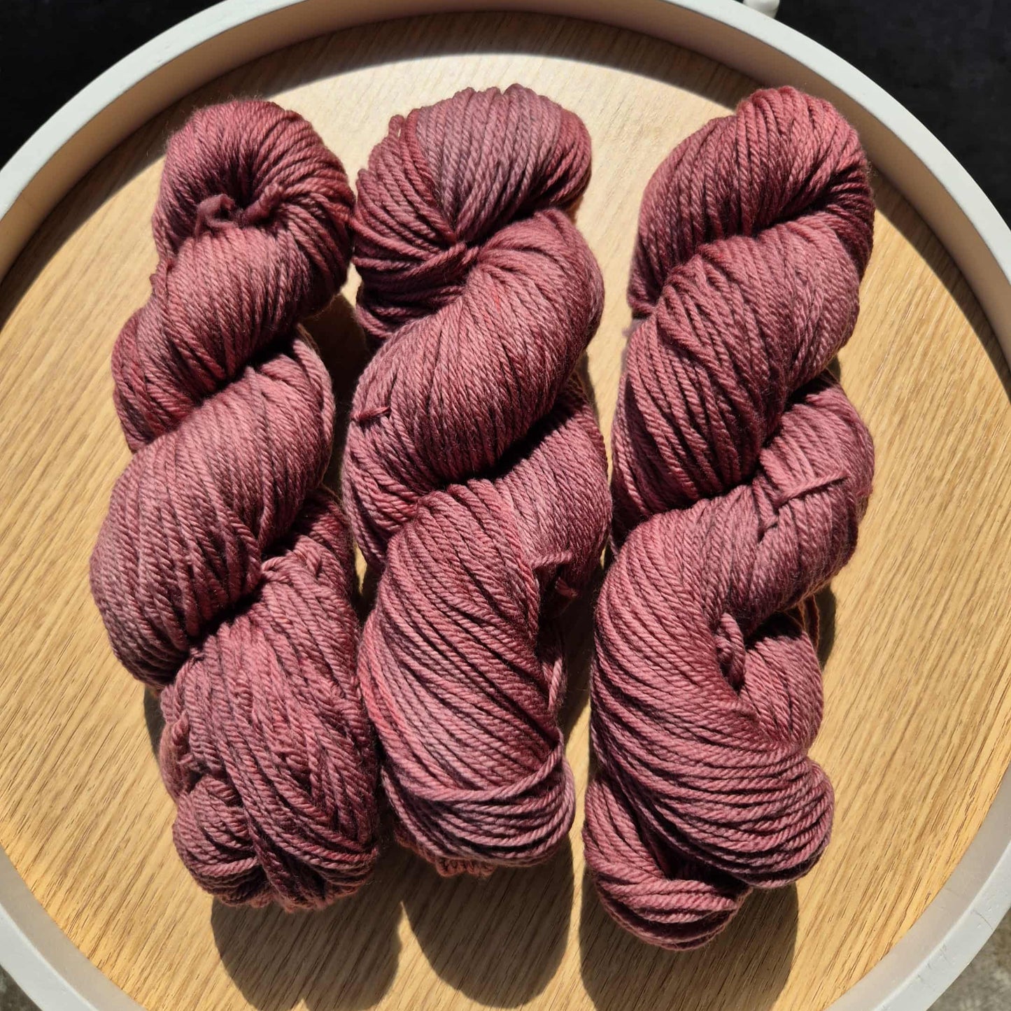 Three skeins of red wine coloured yarn on a wooden board with a white rim.