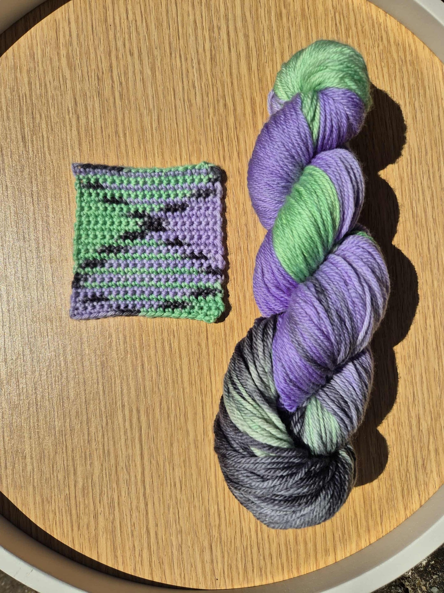 Skein of green, black and purple yarn with a small crocheted swatch on a wooden surface