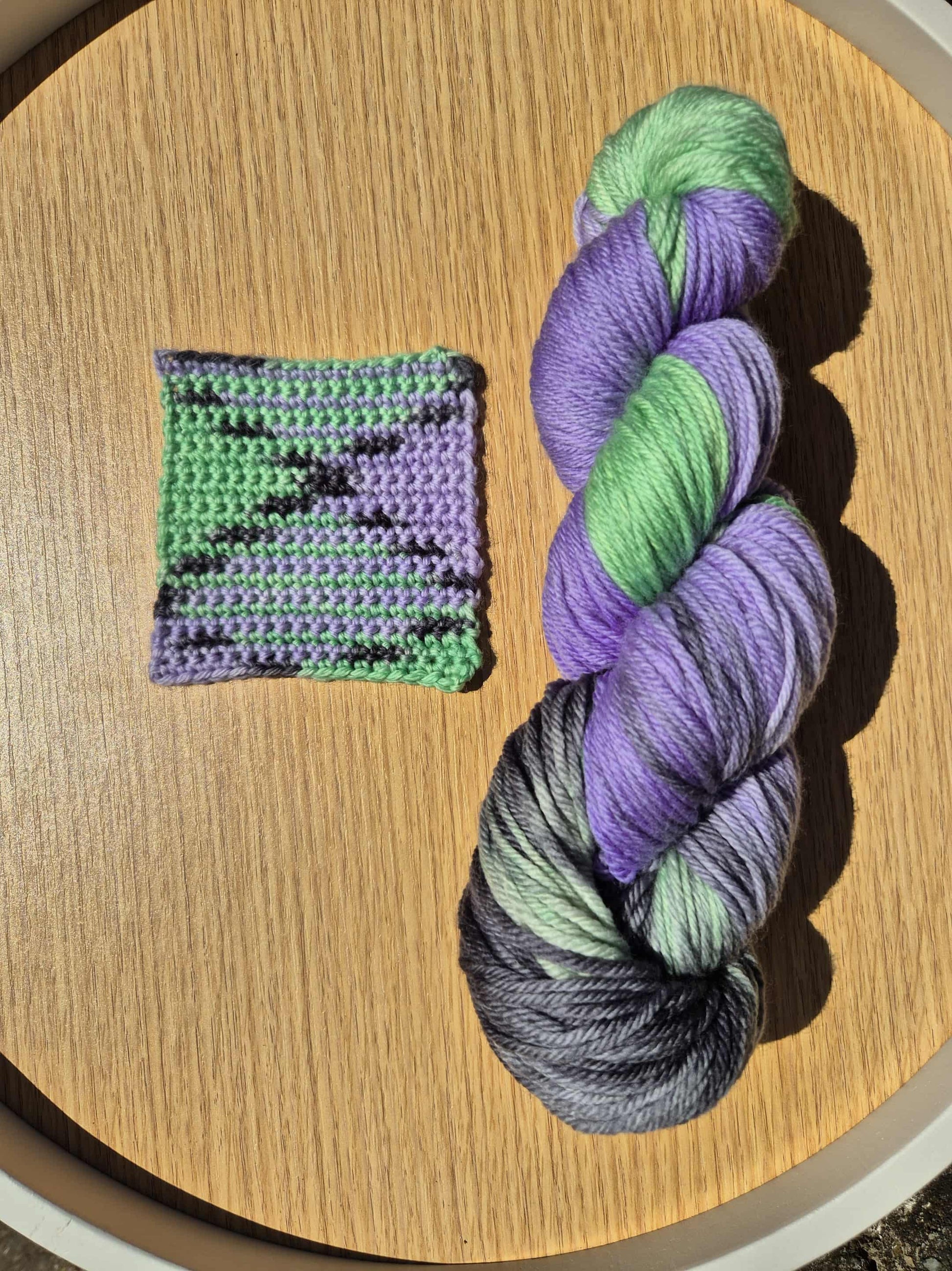 Skein of green, black and purple yarn with a small crocheted swatch on a wooden surface