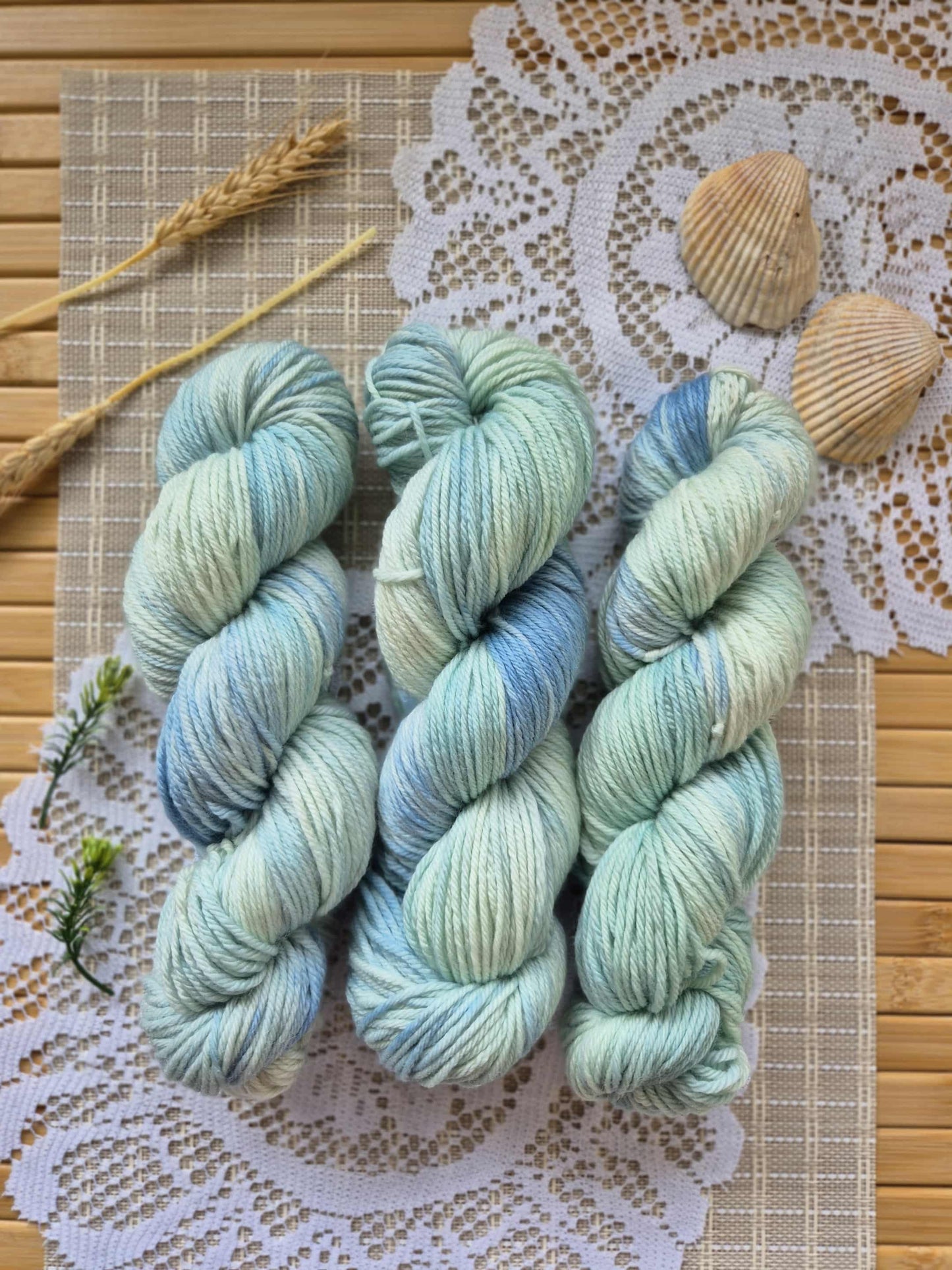 Three skeins of yarn in blue and green on a lace doily with wooden sticks and shells.